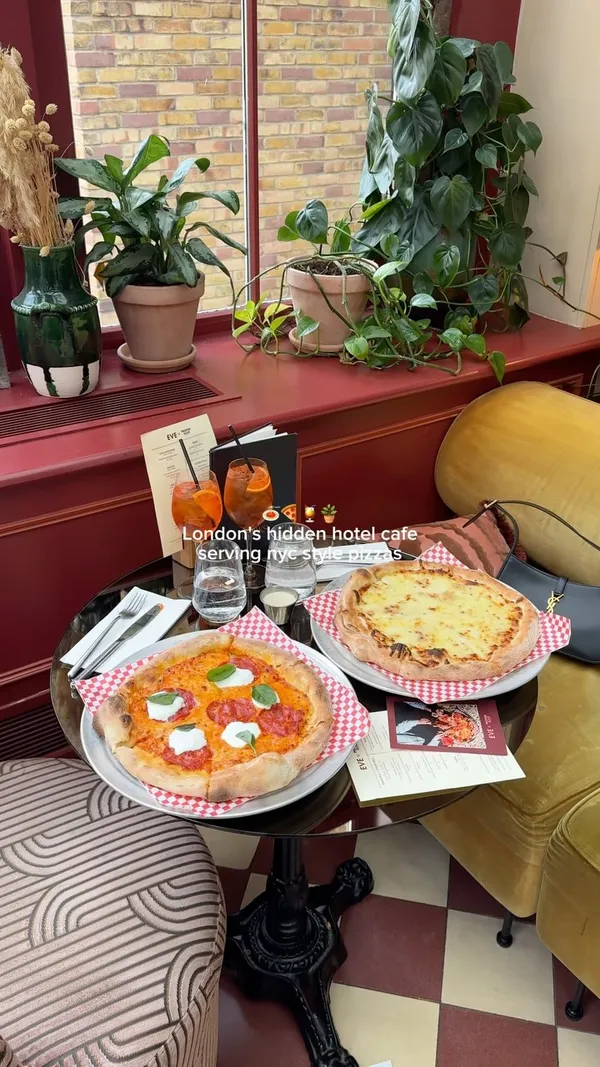 The video showcases the EVE x Proper Slice cafe located within the Ember Locke hotel in Kensington, London. It features the cafe's interior, highlighting its decor and natural light. The video displays various food and drink items, including NYC-style pizzas (one with pepperoni, mozzarella, and basil, and another with cheese), spicy vodka rigatoni, and Aperol spritzes garnished with orange slices. The presentation emphasizes the visual appeal of the food and drinks. The main focus is on showcasing the cafe as a versatile spot for date nights, coffee catch-ups, or solo dining.