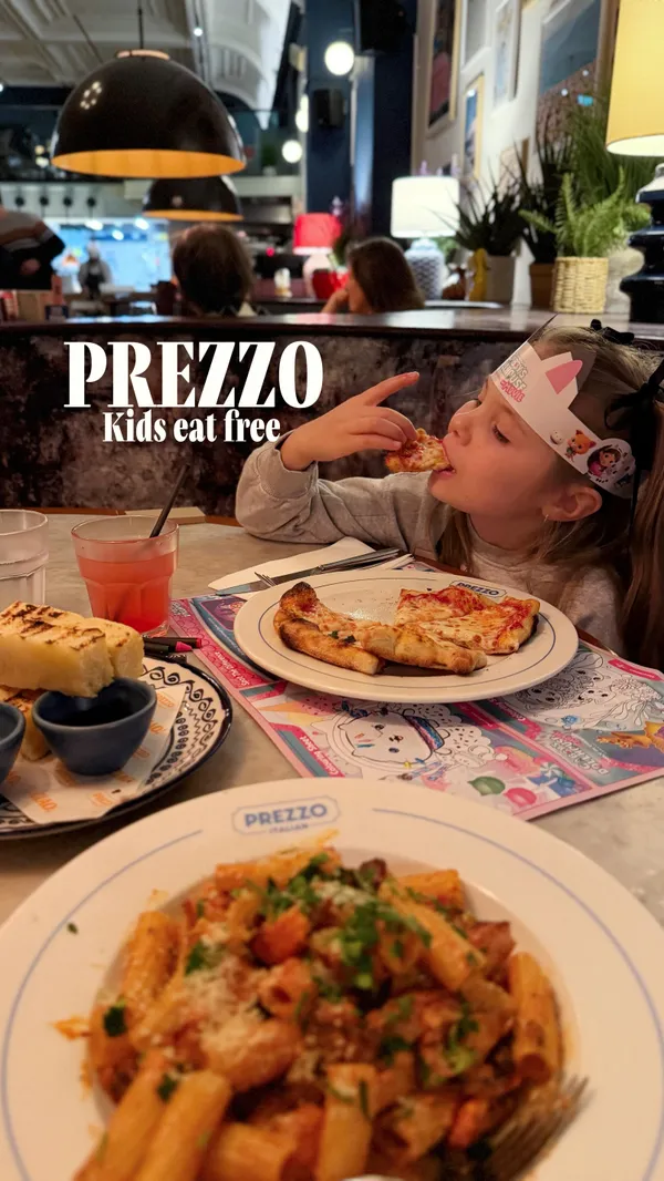 The video promotes a 'Kids Eat Free' offer at Prezzo restaurant, in collaboration with Gabby's Dollhouse. It follows a young girl and her mother during their meal. The video showcases the restaurant's interior, the child enjoying themed activities like coloring, and various food items. Dishes featured include fried shrimp, mozzarella sticks, a cat-shaped pizza, a rigatoni pasta dish, rainbow doughnuts with chocolate sauce, and a Biscoff cheesecake.