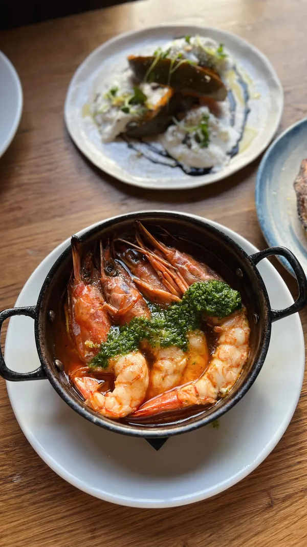 A montage video showcasing a variety of Spanish and Italian tapas dishes from a tasting menu at a London restaurant. The video displays several plates including olives, padron peppers, anchovies in oil, prawns in a garlic sauce, roasted pumpkin with burrata, pan-seared sea bass, pork cheeks with corn, bacon-wrapped skewers, grilled lettuce, and a churro-tiramisu hybrid dessert.