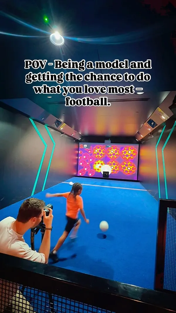 The video shows a person playing an interactive football simulation game at an indoor venue called Toca Social. The player kicks a football towards a large screen displaying targets and scores. Another person is seen filming the player. The video includes a text overlay and background music.