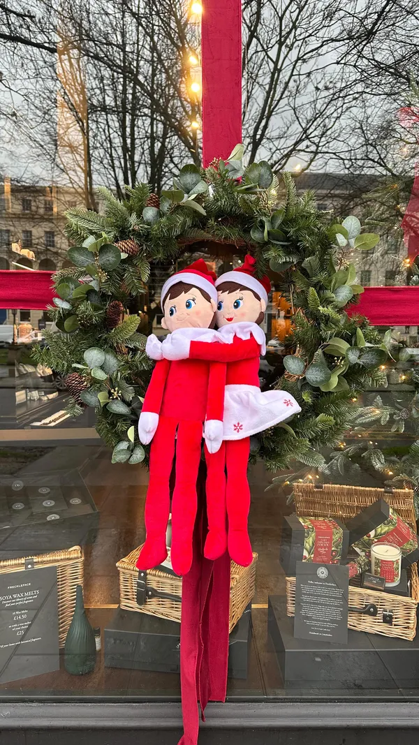 A festive montage video showcasing the Christmas decorations and atmosphere in the town of Cheltenham. The video features a wide array of businesses, including restaurants, cafes, and retail shops, highlighting their decorated window displays for a competition. A recurring theme is the use of 'Elf on the Shelf' dolls placed in various creative settings. The content also includes shots of an ice rink, Christmas market stalls, and large decorative installations within a shopping centre, promoting the town as a festive destination.