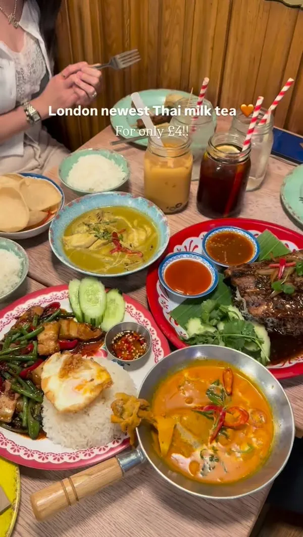 The video showcases a dining experience at a Thai restaurant, Rosa's Thai in London. It features a large spread of various Thai dishes on a wooden table, including green curry, a red curry, stir-fried pork belly with green beans and a fried egg on rice, grilled pork ribs, and a mixed appetizer platter. The video also highlights beverages like Thai milk tea and Thai iced tea served in mason jars. There are close-up shots of the food, a person drinking the milk tea, and a brief view of the restaurant's interior. The main focus is on the food variety and a special offer mentioned in the text overlay.