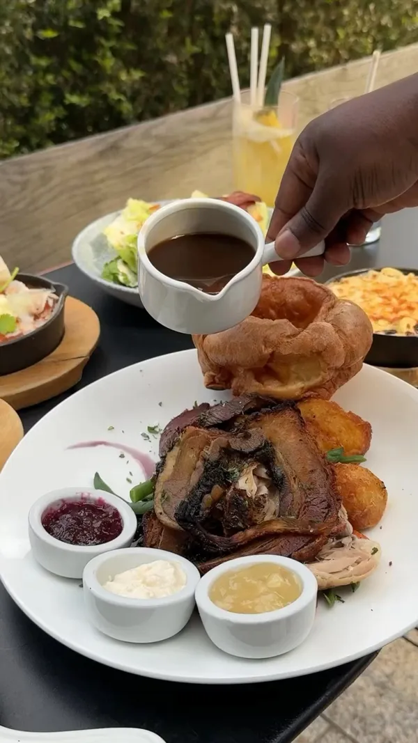 The video showcases No. 17 Dickens Yard, a restaurant in Ealing Broadway, emphasizing its casual modern vibe and patio dining. It features a Sunday roast with beef, chicken, potatoes, and Yorkshire pudding, along with other dishes like a house burger, chicken tenders, and lamb meatballs. The food is presented on white plates with various sauces and condiments, using a mix of overhead and close-up shots to highlight its visual appeal.