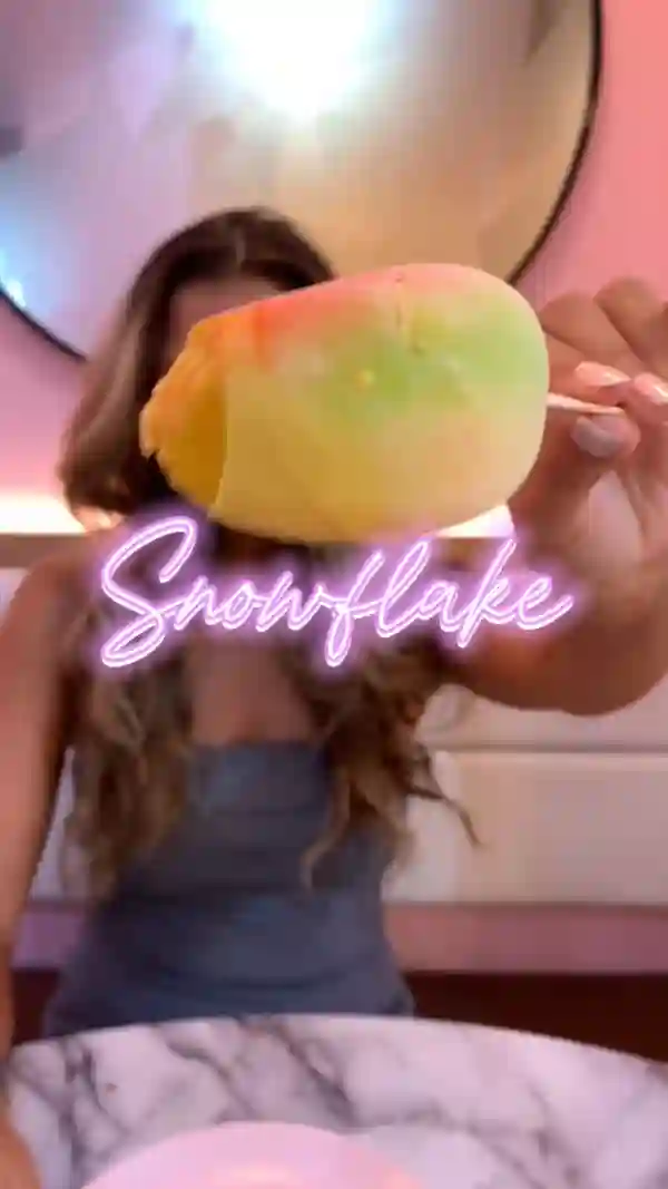 The video showcases a young woman enjoying a mango-flavored ice cream at Snowflake Gelato in London.  The ice cream is vibrant, multi-colored, and appears creamy and delicious. It is presented on a stick in a unique shape. The setting is a modern cafe with pink accents. The main focus is on the ice cream and the woman's positive reaction to it.