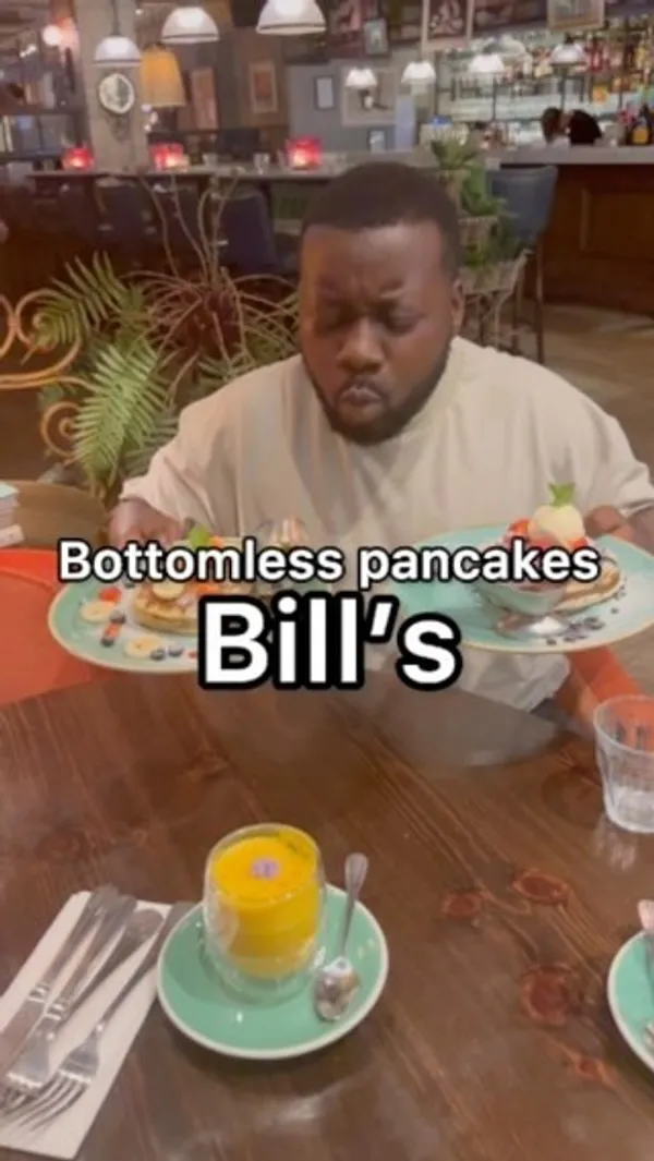 The video showcases Bill's restaurant, focusing on their bottomless pancakes deal for £9.50. It features two pancake options: one with chocolate sauce, chocolate biscuit crumbs, strawberries, and ice cream; the other with fruit (strawberries, bananas, and blueberries) and pancake syrup. Both options are served with unlimited hot drinks (cappuccino, matcha, latte shown). The restaurant's interior is also shown. The main focus is a meal showcase.