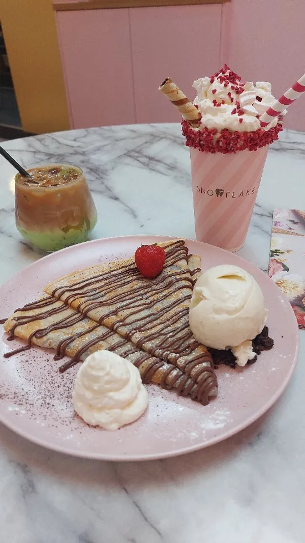 The video showcases Snowflake Gelato in Soho, London. It features crepes with Nutella, whipped cream, and vanilla ice cream; a strawberry and cream sundae shake; and an iced pistachio latte. The presentation is visually appealing, with pastel colors and a focus on the dessert's textures and details.  The video highlights the use of 100% natural ingredients and organic Jersey milk and cream.