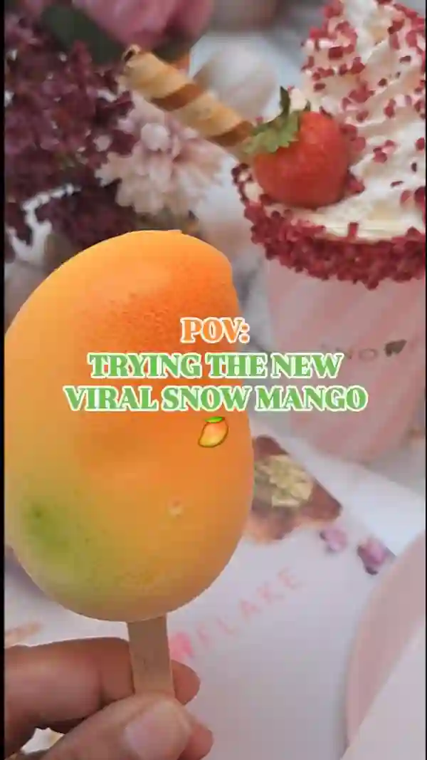 The video showcases a person trying the Snow Mango dessert at Snowflake Luxury Gelato in Soho, London.  The Snow Mango is a mango-shaped dessert, seemingly made with real fruit and white chocolate.  It is presented on a stick. A creamy beverage with whipped cream and red sprinkles is also shown. The video focuses on the visual experience of trying the new dessert, highlighting its unique shape and color.