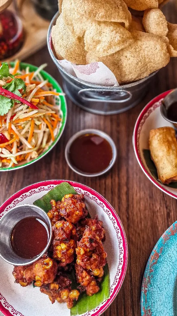 The video begins with an exterior shot of Rosa's Thai restaurant, showing the red facade and outdoor seating. The influencer then enters the restaurant. Inside, there are shots of the interior, including tables, decor, and the kitchen area. Close-ups of various Thai dishes are shown, including papaya salad, spring rolls, sweetcorn fritters, chilli and basil chicken with rice and cucumber, and green curry with rice. There are also shots of drinks being served and the influencer sitting at a table. The video ends with a shot of a banner detailing 'Rosa's Story' and 'Chef Saiphin'.