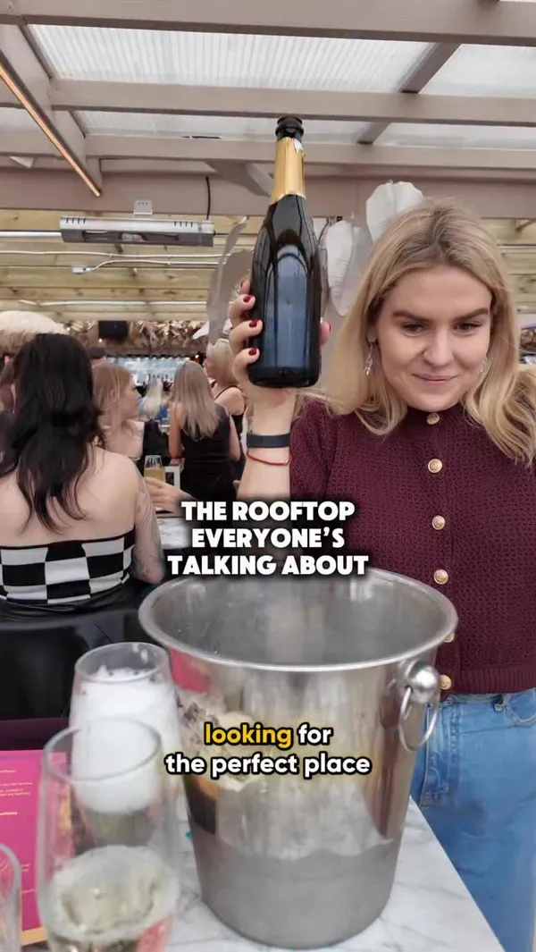 The video showcases a bottomless brunch experience at a rooftop bar called Circe's Rooftop. It features two women enjoying their time, pouring prosecco and cocktails from a jug. The main food item shown is chicken and waffles. The video heavily emphasizes the party atmosphere, with clips of unique performers, including a person covered in silver glitter and another in a black leather outfit and mask, dancing among the tables. There is also a live singer and a DJ. The overall vibe is energetic, fun, and celebratory.