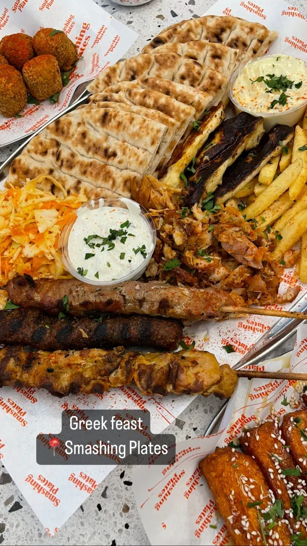 The video showcases a large Greek food platter from Smashing Plates, a restaurant in London. The platter includes grilled meats (souvlaki, chicken, pork), halloumi, pita bread, fries, salad, and various dips.  Additional sides of halloumi fries are also shown. The food is presented on several Smashing Plates branded paper sheets. The video is primarily a close-up food showcase.