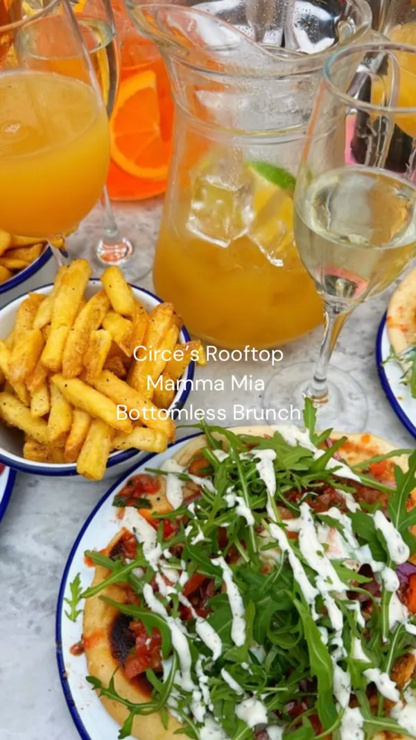 This video showcases a bottomless brunch event at Circe's Rooftop in Waterloo, London. The video features the venue's decor, including straw umbrellas and pink furniture. It shows people enjoying cocktails, prosecco, and wine, as well as food like chicken gyros and fries. The video also highlights the entertainment, including West End singers and a drag queen, with people dancing and having fun. There are also shots of people toasting with drinks and close-ups of the food.