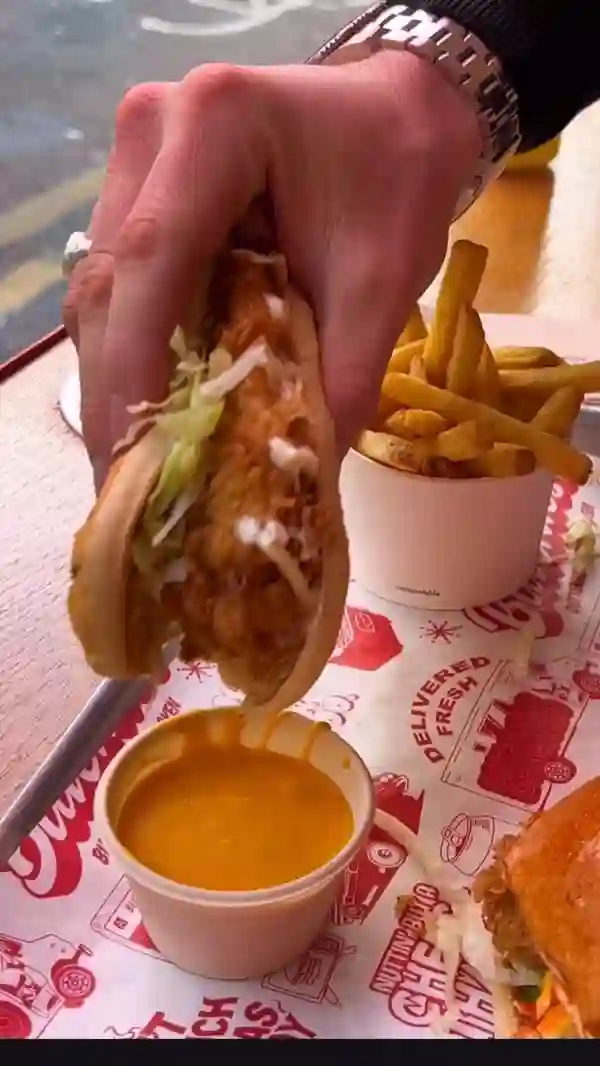 The video showcases a new menu item from Butchies London, a chicken dawg. The setting appears to be an outdoor seating area of a restaurant. The chicken dawg consists of chicken tender, mayo, and lettuce in a brioche roll. It's dipped in a cheese sauce, and a side of fries is visible. The presentation highlights the gooey cheese pulling away from the sandwich. No dietary information is explicitly mentioned.