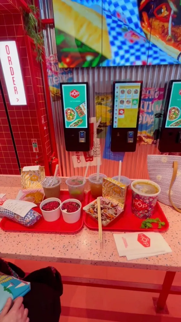 This video showcases Hop Vietnamese restaurant located at Bond Street station in London. The video starts with a touchscreen kiosk for placing orders, proceeds to show the overall ambiance of the restaurant including bright lights and hanging lanterns. Then, it shows the process of ordering and receiving the food, which includes chicken Pho, Viet slaw, and drinks like lemonade and bubble tea. Finally, it shows the food and drinks being enjoyed in the restaurant, with additional shots of the restaurant's interior design. The video has a vibrant and energetic feel throughout.