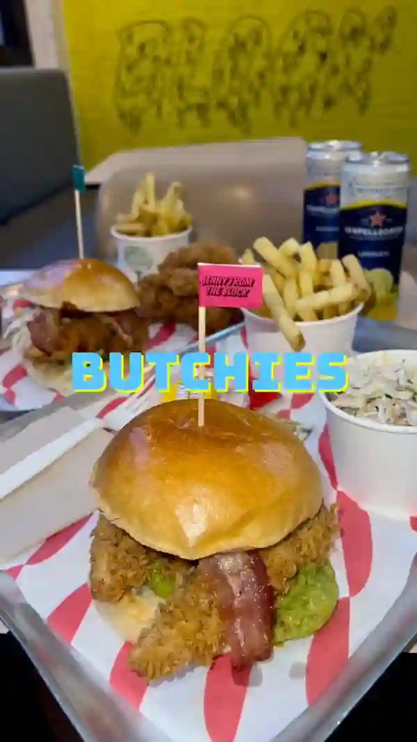 The video showcases a meal at Butchies in Shoreditch. It features chicken sandwiches with bacon and guacamole, crispy chicken tenders, fries, coleslaw, buffalo sauce, and Sanpellegrino Limonata. The food is presented on a red and white striped tray. The primary focus is on the enjoyment of the meal, emphasizing the deliciousness of the food and the pleasant dining experience.