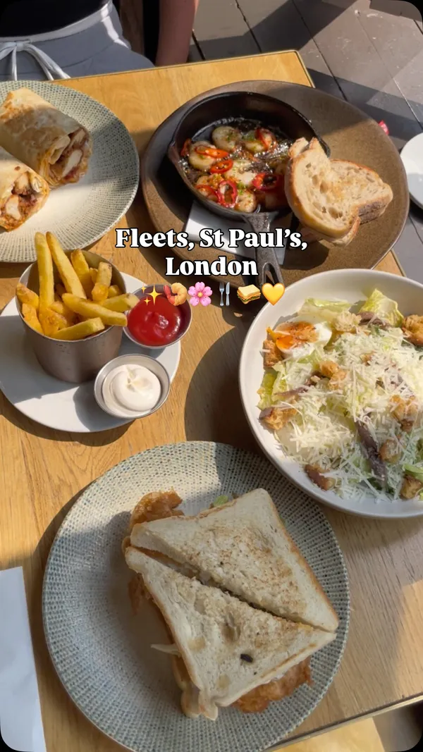 This video showcases a lunch experience at Fleets, a restaurant and bar located near St Paul's in London. The video highlights their '10 for £10' lunch menu. The food items featured include sizzling chili and garlic king prawns with sourdough bread, a large haddock fish finger sandwich, a Caesar salad with anchovies and a soft-boiled egg, and a crispy chicken wrap. The video also shows cocktails, specifically a Hugo Spritz and a Pornstar Martini. The interior of the restaurant is shown, featuring a large, well-stocked bar, a neon 'Fleets' sign, and decorative pink cherry blossom trees.
