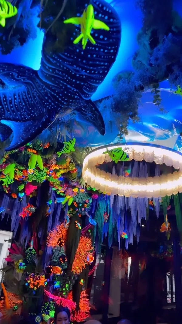 The video showcases Arc Le Salon, a Mayfair lounge with an "Under the Sea" theme. The decor features glowing UV lighting, artificial coral, and marine life decorations. The food and drink items featured are: Rose Harrisa Hummus, Mushroom & Truffle Croquettes, Crispy Tempura Prawns, Signature Beef Burger, Limousin Sirloin Steak, Waffle Delight, Pink Beryl cocktail, and Fire and Magic cocktail. The food is presented attractively on elegant plates. Dietary information includes a mention of Halal options and that alcohol is served. The main focus is showcasing the atmosphere and food of the lounge.