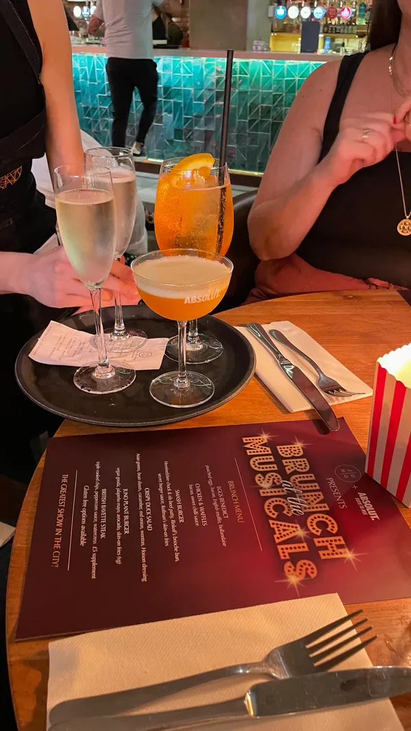 The video showcases a musical theatre-themed bottomless brunch at Juno Rooms, a bar in London. It features various food items, including a smash burger with fries and several cocktails and drinks (bottomless Prosecco and cocktails). The food is presented attractively, and the video highlights the lively atmosphere and drag performances. Dietary information isn't explicitly shown aside from the mention of bottomless options.