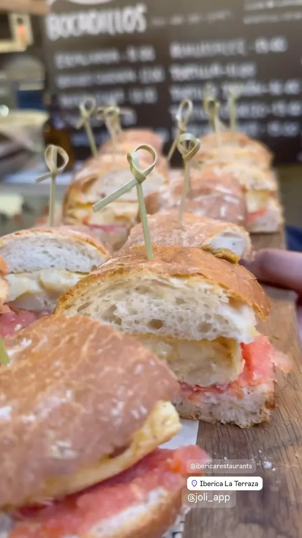 The video showcases the reopening party of Iberica La Terraza in Canary Wharf. It features various food items, including small sandwiches with ham and egg, sliced ham, and other tapas-style dishes.  The drinks shown include beer. The setting is an outdoor terrace with a modern look, and the food presentation is simple, focusing on showcasing the food itself. No dietary information is explicitly stated.