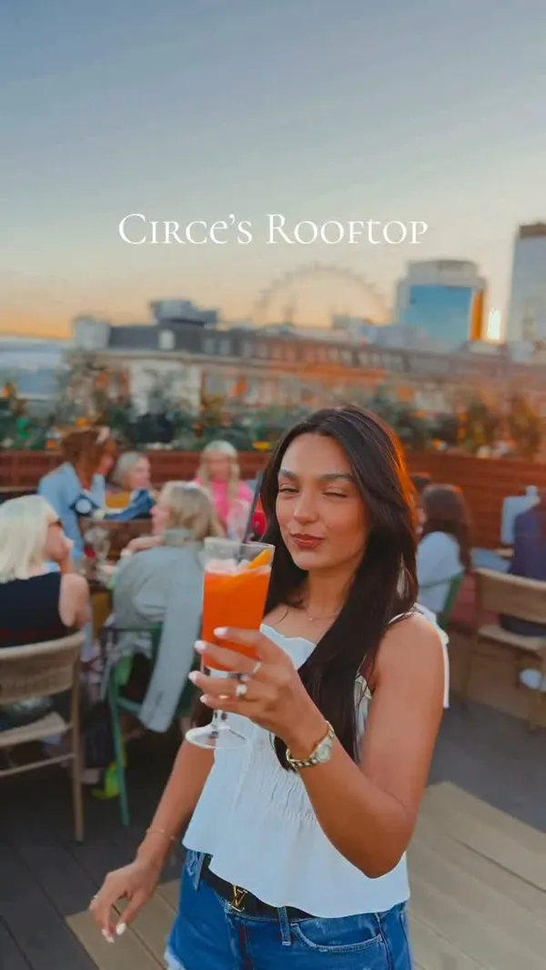 A short, fast-paced video montage showcasing Circe's Rooftop bar in London. It features a woman holding various cocktails (an orange spritz, piña colada-style drinks, a coupe cocktail) against the backdrop of the London skyline at sunset. Shots of food are included, such as skewers with salad and a lettuce wrap dish. The video highlights the atmosphere with clips of a DJ, the bar area, other patrons, and the scenic views. Text overlays label the location, food, drinks, and include a call to action to book a table.