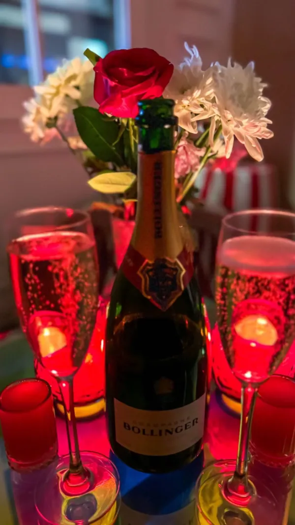 The video showcases a romantic Valentine's Day date night setup at The Little Scarlet Door.  The scene features two champagne flutes filled with Bollinger champagne, a bottle of Bollinger champagne, and a floral arrangement with red roses and white flowers. Red candlelight illuminates the scene, which is set on a glass table.  The video includes a short clip of someone pouring more champagne into a flute.