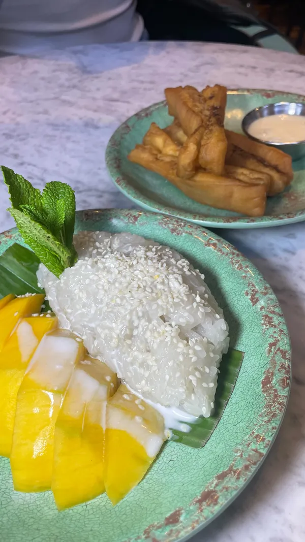 The video documents a dining experience at a Thai restaurant. It features a bubble tea drink, a large sharing platter with various fried items (spring rolls, squid, chicken wings, satay) served with multiple dipping sauces and prawn crackers. It also showcases individual dishes including Larb (minced meat salad) served in lettuce cups, green curry with chicken and bamboo shoots, steamed rice, grilled eggplant topped with herbs and soft-boiled eggs, and mango sticky rice. The video includes shots of someone stirring the bubble tea, dipping a spring roll into sauce, pouring sauce over the satay, and eating the mango sticky rice and fried banana fritters.