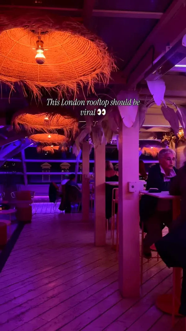 A fast-paced video montage of a London rooftop bar. The video showcases the venue's interior with purple and orange lighting, outdoor seating with futuristic-looking igloos overlooking the city at night (including the London Eye), a bartender making and pouring an orange cocktail, and various food dishes. The food includes a passionfruit martini, another cocktail with a dried apple slice, hummus with pita bread, falafel, a fish dish, and sliced steak being drizzled with a sauce. The video ends with a shot of the lively crowd inside the bar.