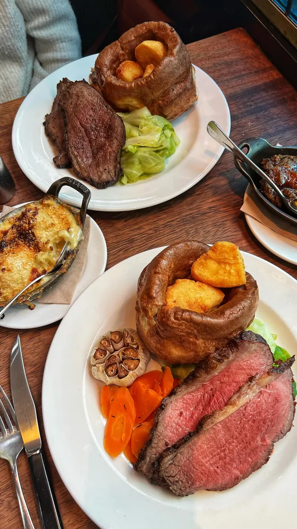 The video showcases a Sunday roast at Hawksmoor Spitalfields in London.  The main focus is on the pouring of gravy over a sliced roast beef, accompanied by Yorkshire puddings, roasted vegetables (carrots and garlic), and what appears to be cauliflower cheese in the background. The presentation emphasizes the rich color and texture of the meat and gravy.