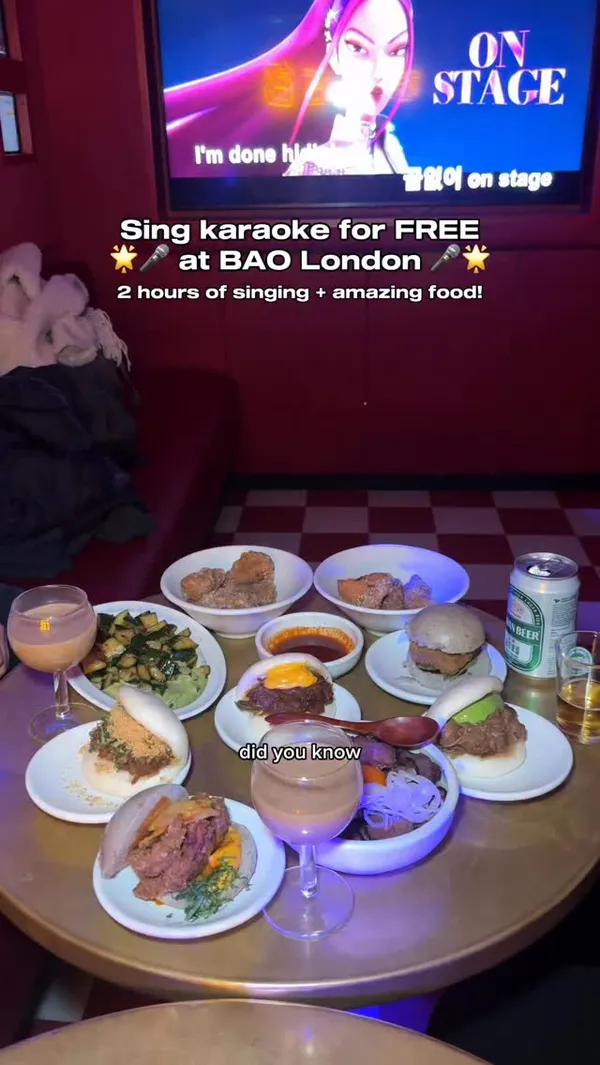 The video promotes a free karaoke experience at BAO London. It shows a group of friends enjoying a private karaoke room, singing and playing tambourines. The video features a wide variety of food items, including several types of bao buns (with fried chicken, pulled beef, and other fillings), a beef and egg rice bowl, fried chicken pieces, and a cucumber salad. The group is also shown toasting with drinks. The overall atmosphere is fun, energetic, and social.
