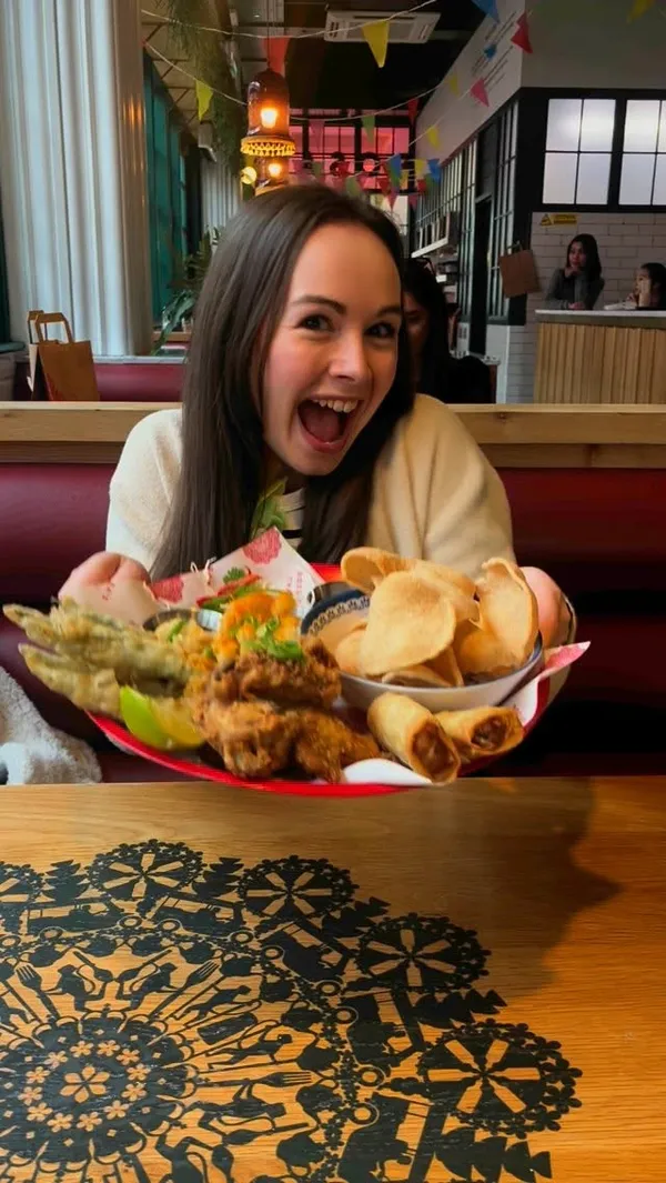The video features a woman and a man dining at a Thai restaurant. It showcases a large sharing platter with various starters including fried chicken, spring rolls, calamari, and prawn crackers with dipping sauces. Main courses shown are Pad Thai with prawns and a Chilli & Basil Stir Fry with minced chicken, rice, and a fried egg. A dessert of a brownie with ice cream and chocolate sauce is also featured. The video includes shots of the diners interacting with the food, such as dipping spring rolls and eating Pad Thai.