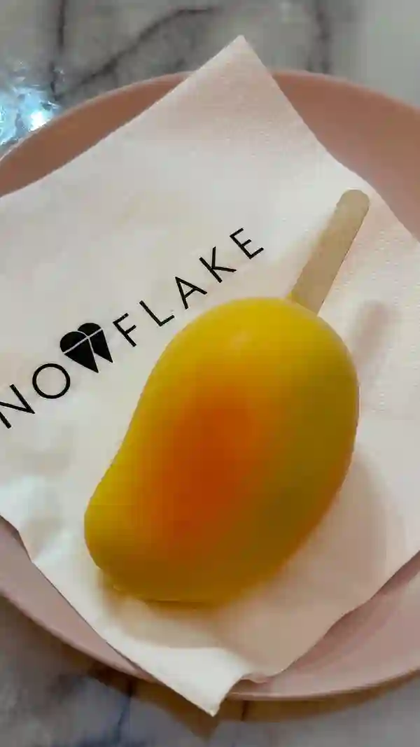 The video showcases a young woman enjoying a mango-shaped ice cream on a stick at Snowflake Gelato in London. The setting is a pink-themed cafe with marble tables.  The ice cream is yellow and orange, presented on a stick. The video focuses on the woman's experience of eating the ice cream.