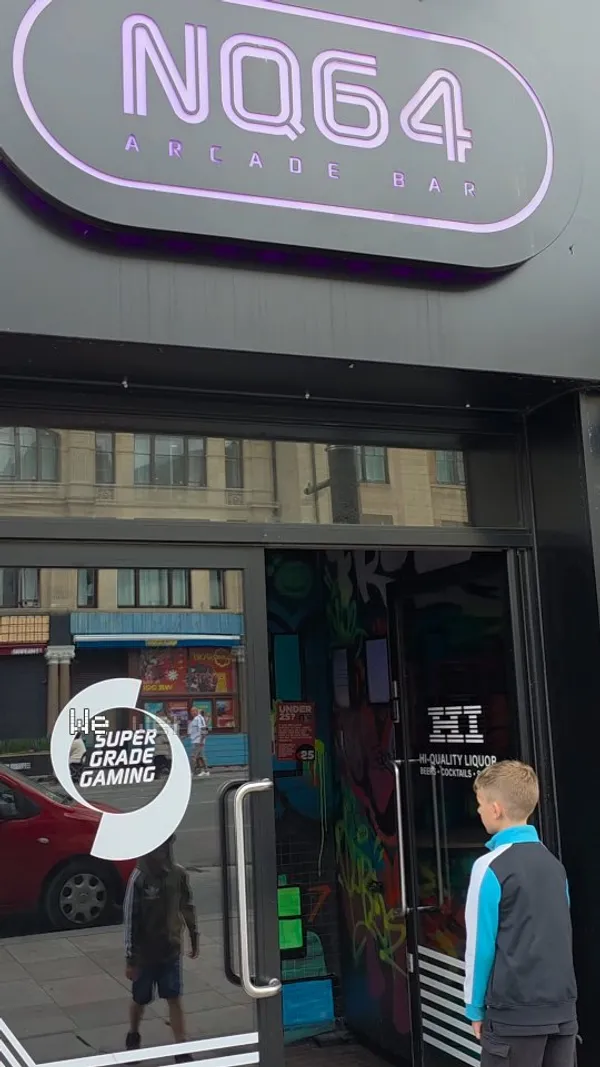 The video promotes a family-friendly event at NQ64, a retro arcade bar. It begins with an exterior shot before moving inside to showcase the neon-lit, graffiti-covered interior. The main focus is on children playing various classic arcade games, including Bubble Bobble, a light gun shooter, Pac-Man, Mario Kart, and The Simpsons. The video also briefly shows the well-stocked bar area, indicating it's a licensed venue. The overall theme is retro gaming in a vibrant, visually stimulating environment.