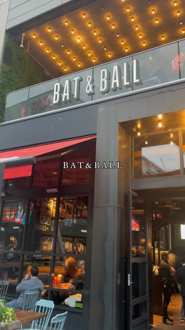 The video showcases Bat & Ball, a bar with a dartboard. It features two pizzas (one with meat, the other with chicken and vegetables), a burger, and a cocktail. The food is presented on wooden tables. The primary focus is on the venue and the food and drink options.