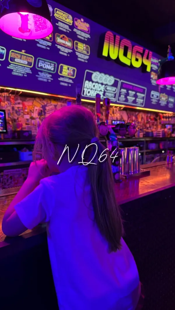 The video promotes a family-friendly event at the NQ64 arcade bar. It showcases the venue's dark, neon-lit, and graffiti-art interior. The content features a family (parents and a child) playing a wide variety of retro arcade games, including Pac-Man, Time Crisis II, basketball hoops, a dance machine, and an Avengers pinball machine. The video also includes shots of a bartender preparing a pink cocktail.