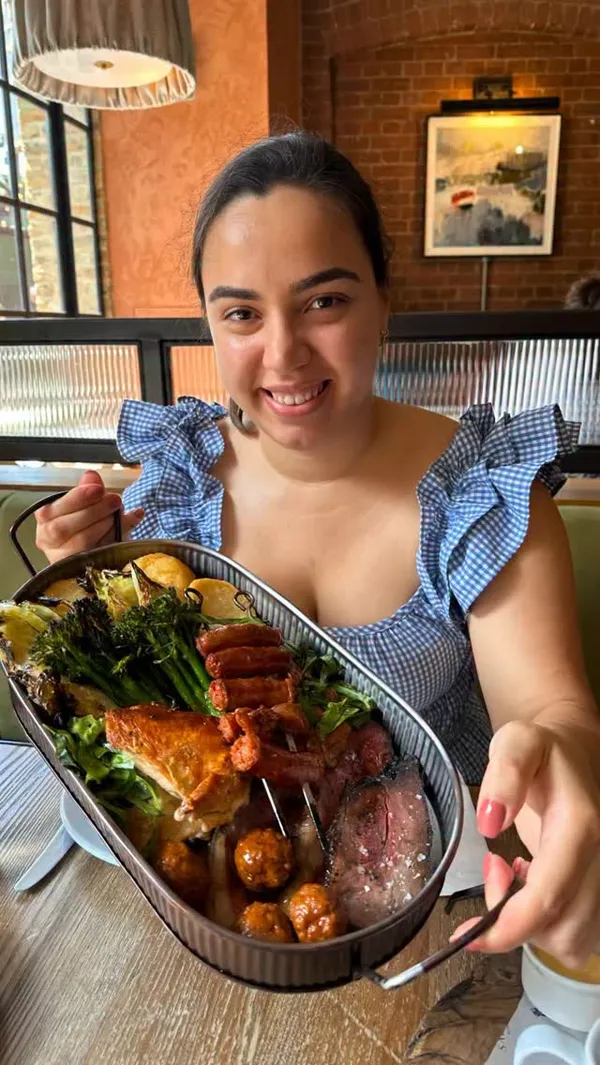 The video promotes a Spanish-Italian style Sunday roast from Salt Yard, a restaurant located near Borough Market in London. It showcases a large sharing platter containing various meats like crispy pork, beef, chicken, sausages, and meatballs, alongside roasted vegetables such as potatoes, cabbage, and broccoli. A key visual is a rich gravy being poured over the food. The video also provides glimpses of the restaurant's rustic and cozy interior, featuring brick walls and an open kitchen.