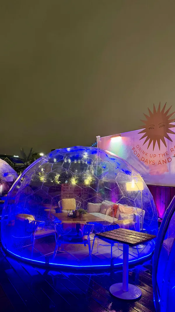 The video showcases a stylish rooftop bar in London called Circe's Rooftop. The setting features a modern interior with a glass roof and an outdoor terrace with heated, transparent domes, all illuminated by vibrant pink and blue neon lights. The content highlights the preparation and serving of colorful cocktails and a variety of small plates, including hummus, fried chicken, lamb kofta, and prawns on toast. The main focus is on the venue's unique ambiance, its special 'Witching Hour' deal, and the overall experience of socializing with food and drinks.