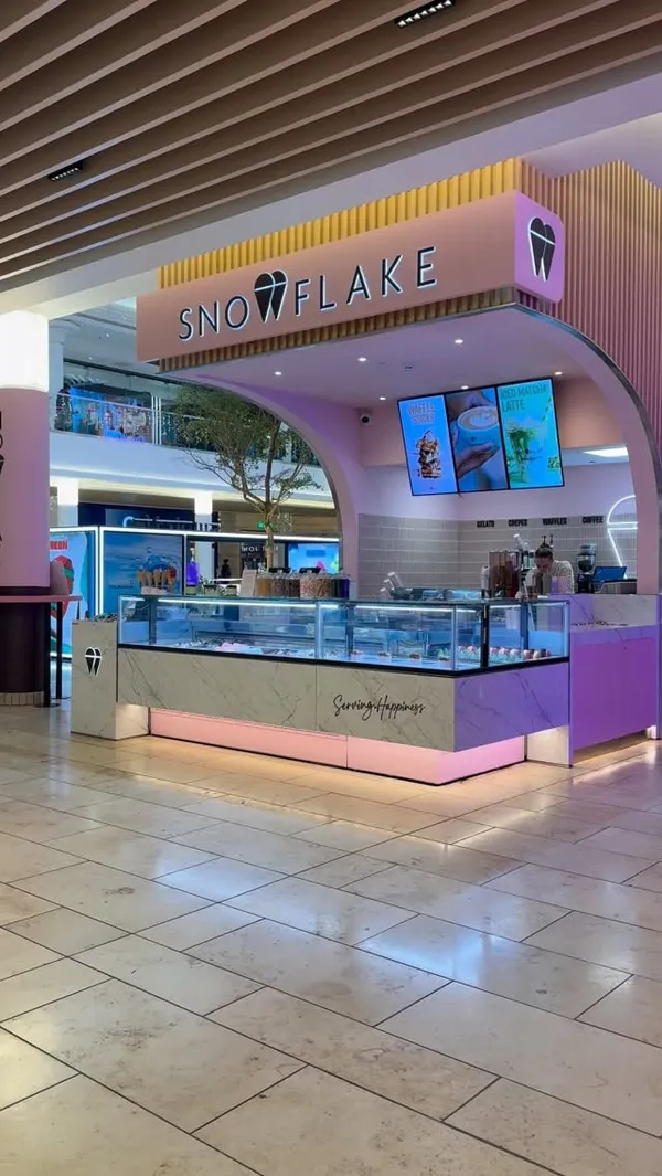 The video is a showcase of a "Snowflake" gelato kiosk located in a shopping mall. It begins with a wide shot of the storefront, then moves to a close-up of the various gelato flavors in the display counter. The camera pans across the menu, showing items like gelato, crepes, waffles, and drinks. It then highlights festive-themed popsicles, including Christmas tree and Santa shapes. The video also features close-ups of hot chocolate cups with whipped cream and a final shot of waffles on sticks in the display.
