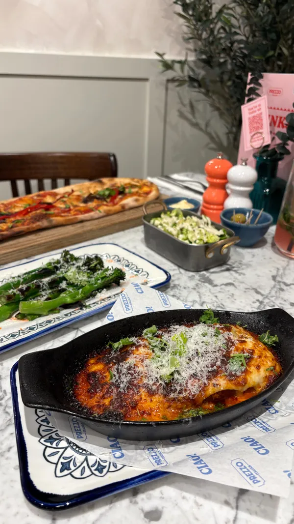 The video showcases a meal at the Italian restaurant chain, Prezzo. It features a variety of dishes, starting with appetizers like Ham & Mozzarella Pastacini, olives, burrata salad, and garlic bread. The main courses shown are a bubbling Lasagne Al Forno and a long Chicken & Roasted Pepper pizza. The video also includes shots of cocktails and side dishes. The content highlights the food being served and eaten, as well as the restaurant's newly refurbished, modern interior, creating a comprehensive overview of the dining experience.