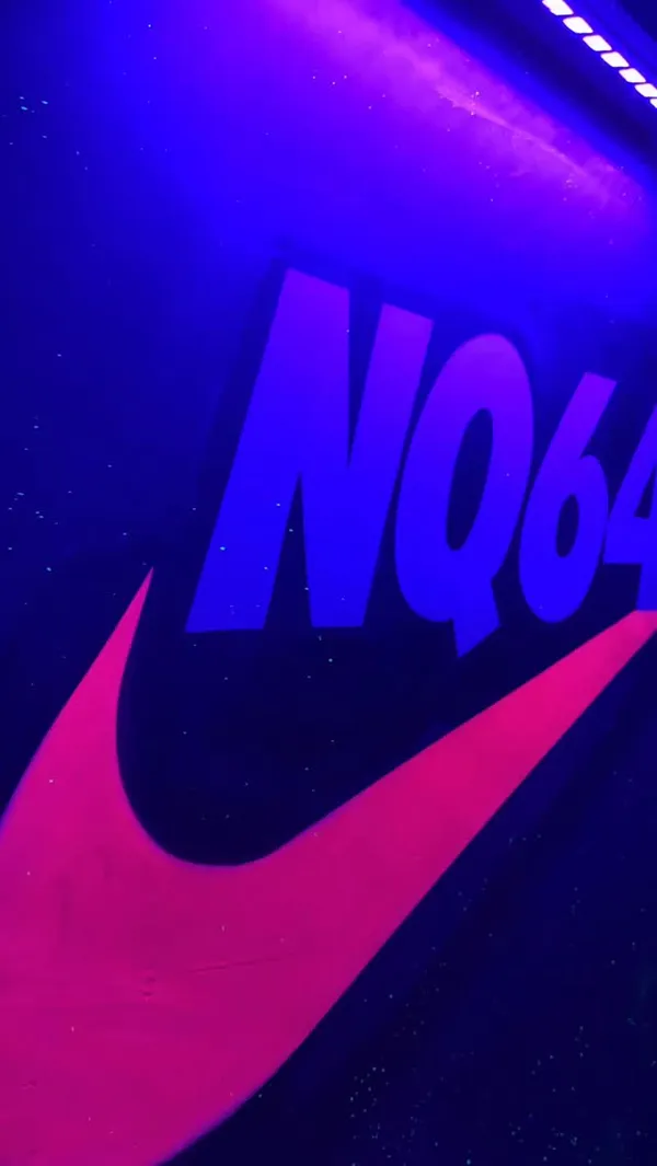 A fast-paced promotional video for NQ64, a retro arcade bar in Manchester. The video showcases the bar's neon and UV-lit interior, featuring its logo and exterior sign. It highlights the drinks, showing a bartender preparing a cocktail with a foam top and close-ups of two different themed cocktails. Various classic arcade games are shown, including Mario Kart, Track & Field, The Simpsons, and a pinball machine. The video captures the lively atmosphere with shots of patrons enjoying the games and drinks.