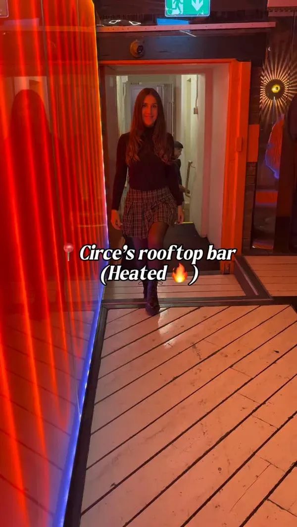 The video showcases Circe's, a heated rooftop bar in London. It begins with a woman entering the venue, followed by shots of the London skyline at night, including The Shard. The video then displays several small plates of food, including scallops in a shell, halloumi fries, calamari with a dip, and caprese skewers. A bartender is shown preparing an Aperol Spritz-style cocktail. The interior decor is highlighted, featuring a disco ball, pink walls with cloud murals, and neon lighting. The video concludes with shots of the lively atmosphere, showing other patrons, the bar area, a DJ, and more views of the city, including the London Eye.