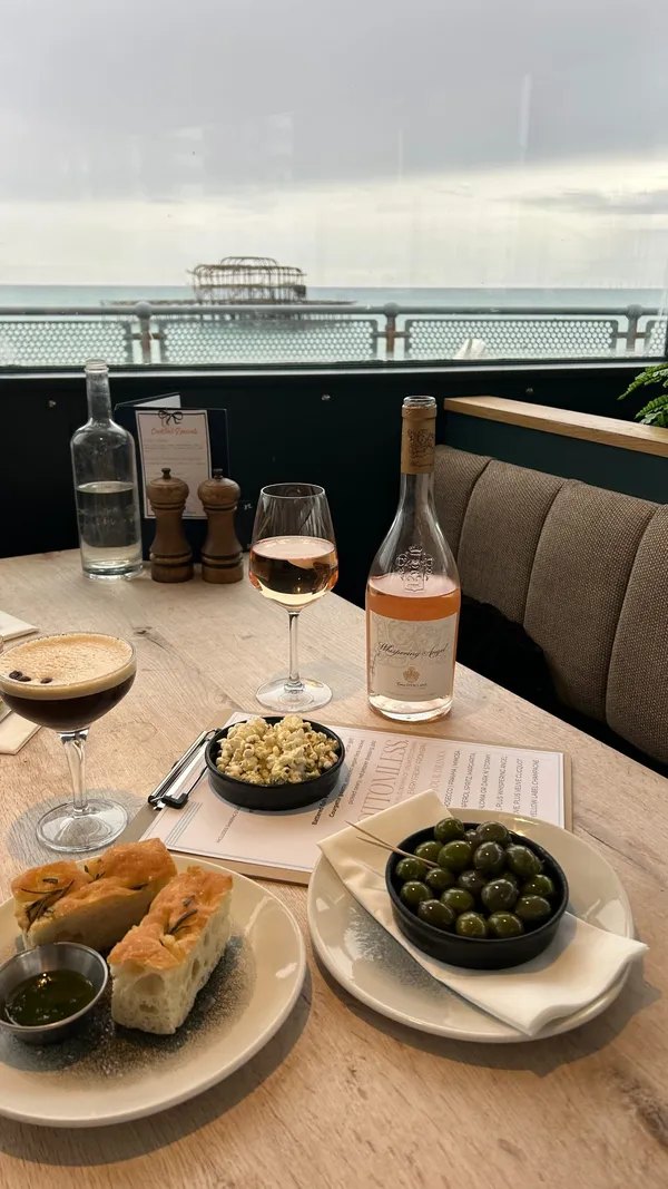 The video promotes a 'Whispering Angel Bottomless Dinner' at The Seahorse restaurant in Brighton. It showcases the restaurant's modern interior, its scenic view of the sea and the derelict West Pier, and the food and drinks included in the offer. Food items shown include olives, focaccia bread, and savoury popcorn as starters, followed by mains of a flat iron steak with fries and a chicken dish. Desserts, including a chocolate fondant and a panna cotta, are also shown but noted as not included in the deal. Drinks featured are Whispering Angel rosé wine, an espresso martini, and other cocktails. The video uses text overlays to detail the different price packages for the bottomless dinner.