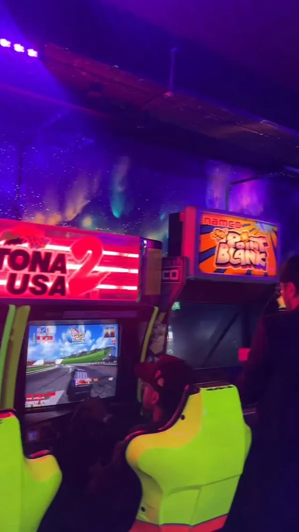 A video showcasing a visit to the NQ64 arcade bar in Soho, London. The video follows two friends as they experience the venue, which features a dark, neon-lit, and graffiti-covered interior. They are shown playing a variety of retro arcade games, including a racing game, Guitar Hero, and Street Fighter, as well as console games like Crash Bandicoot on a PlayStation 1. The video also highlights the bar's cocktails, showing a bartender preparing a 'Kong Island Iced Tea' and the creator enjoying it and another punch-style drink.