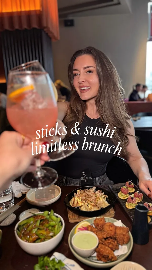 The video promotes a limitless sushi brunch at Sticks'n'Sushi. It showcases a wide array of dishes served, starting with multiple starters like edamame, fried chicken, beef tataki, gyoza, and tuna tartare. This is followed by a platter of assorted sushi rolls and nigiri, and then grilled skewers ('sticks') of salmon and pork. The experience is rounded off with cocktails, wine, and a mango sticky rice dessert. The creator is featured enjoying the meal, and the video also includes shots of the restaurant's modern interior and bar.