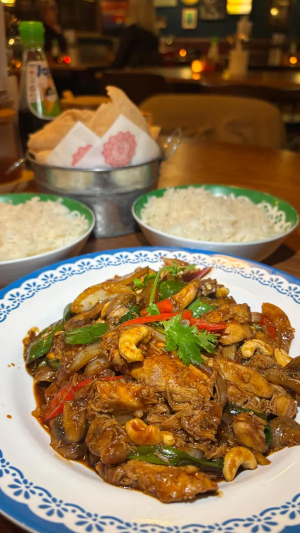 A video montage showcasing a festive meal at Rosa's Thai restaurant. It begins with a close-up of a spicy cocktail, followed by various dishes including a sharing platter with chicken satay and spring rolls, a Thai red curry, and a cashew stir-fry. The video also shows people enjoying the meal, pulling a Christmas cracker, and toasting with shots. Desserts, including a banoffee roti with ice cream, are featured. The video concludes with a gift exchange.