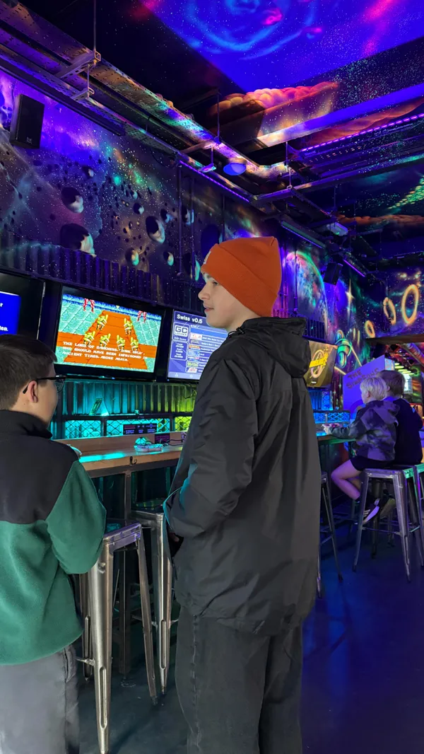 The video promotes a 'Shorty Sundays' family-friendly event at the NQ64 arcade bar in Cardiff. It showcases the venue's exterior and its interior, which is characterized by dark, neon lighting and graffiti art. The video features a variety of retro arcade games being played by a family, including Mortal Kombat, The Simpsons, racing simulators, a Godzilla pinball machine, and Guitar Hero. A child is also shown having a drink at a table.