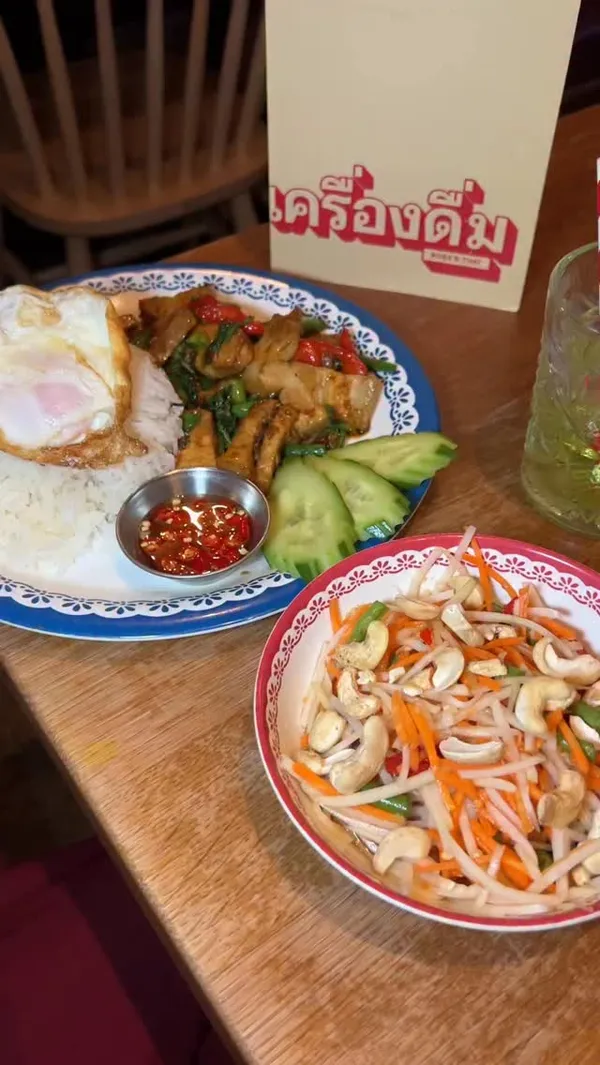 A video review of a meal at Rosa's Thai restaurant. The video showcases several dishes and drinks, including Thai prawn crackers with satay sauce, chili oils, a pork belly pad krapow with rice and a fried egg, and a som tam (papaya salad) with cashews. The drinks featured are a green Melon Basil Smash and a pink Lychee Yuzu Soda. The creator provides a voiceover review of their experience and the food, and concludes by showing the restaurant's menu.