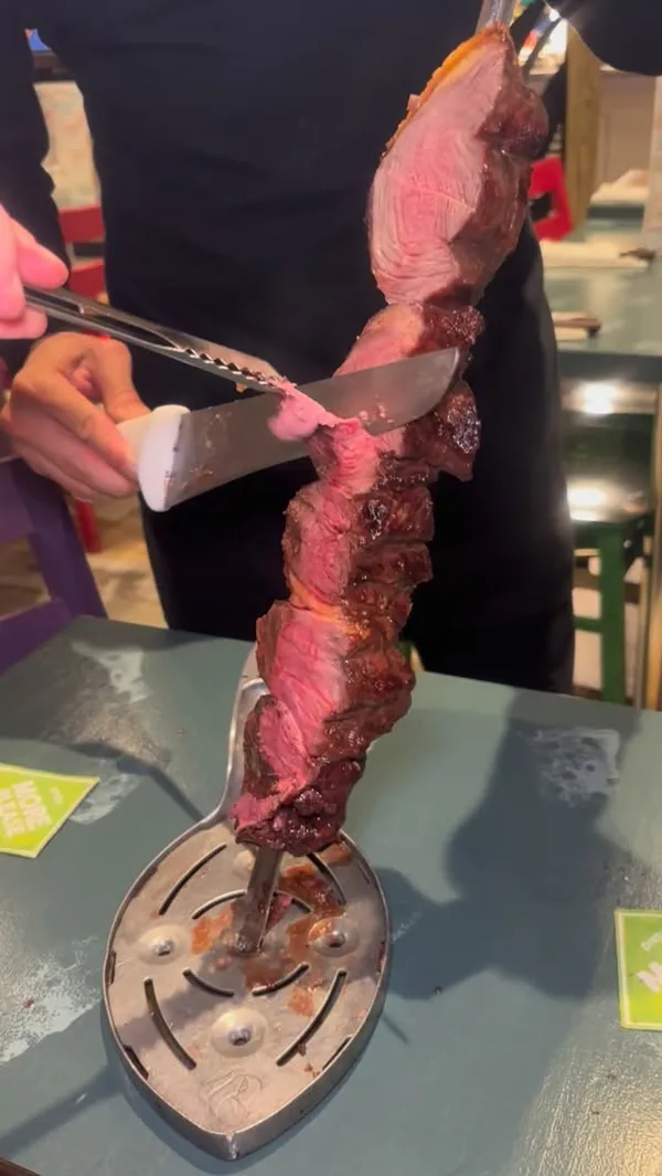 The video showcases the dining experience at Preto, a Brazilian rodizio restaurant. It features a server carving various types of grilled meats, such as steak, chicken, and sausages, from large skewers directly at the customer's table. The video also highlights the restaurant's system for requesting more food using green ("More Please") and red ("I'm Full") cards. It shows a buffet-style salad bar with a variety of fresh salads, traditional Brazilian hot dishes like cheese bread, mashed potatoes, and fries. The video concludes with shots of Caipirinha cocktails.