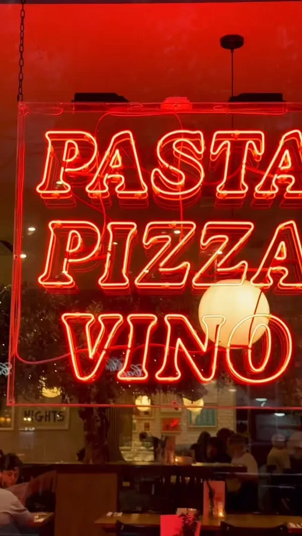 The video showcases the interior and exterior of Prezzo, an Italian restaurant. It begins with a shot of a red neon sign reading "PASTA PIZZA VINO," followed by views of the cozy, warmly lit dining area. The main focus is on the food, featuring several dishes: Italian meatballs in tomato sauce with focaccia, crispy calamari with a dip, a ham and mushroom pizza, spaghetti with king prawns, and crab and lobster ravioli topped with rocket. Action shots include cutting into a meatball, sprinkling cheese on the ravioli, and twirling spaghetti on a fork.