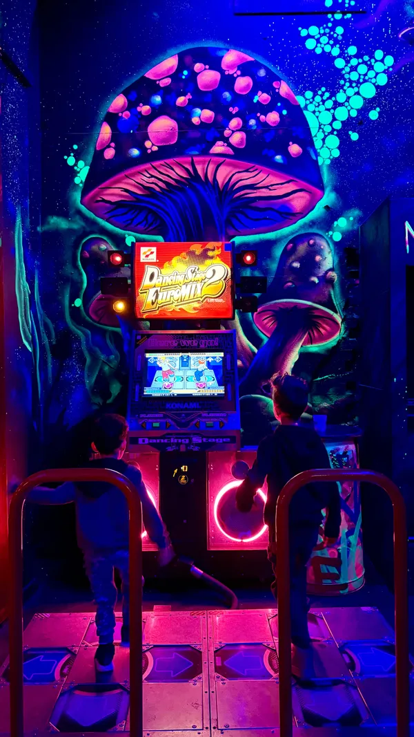 A montage video showcasing a family's visit to the NQ64 retro arcade bar in Nottingham. The video features children and adults playing various classic arcade games, including Dance Dance Revolution, Donkey Kong, Sega Rally, basketball hoops, The Simpsons, pinball, Street Fighter II, Space Invaders, and Mario Kart. It also shows a bartender preparing colorful drinks and the family enjoying them. The setting is a dark arcade with vibrant neon and UV lighting, creating a retro, glowing atmosphere. The main focus is on the variety of games available and the fun, family-friendly experience.