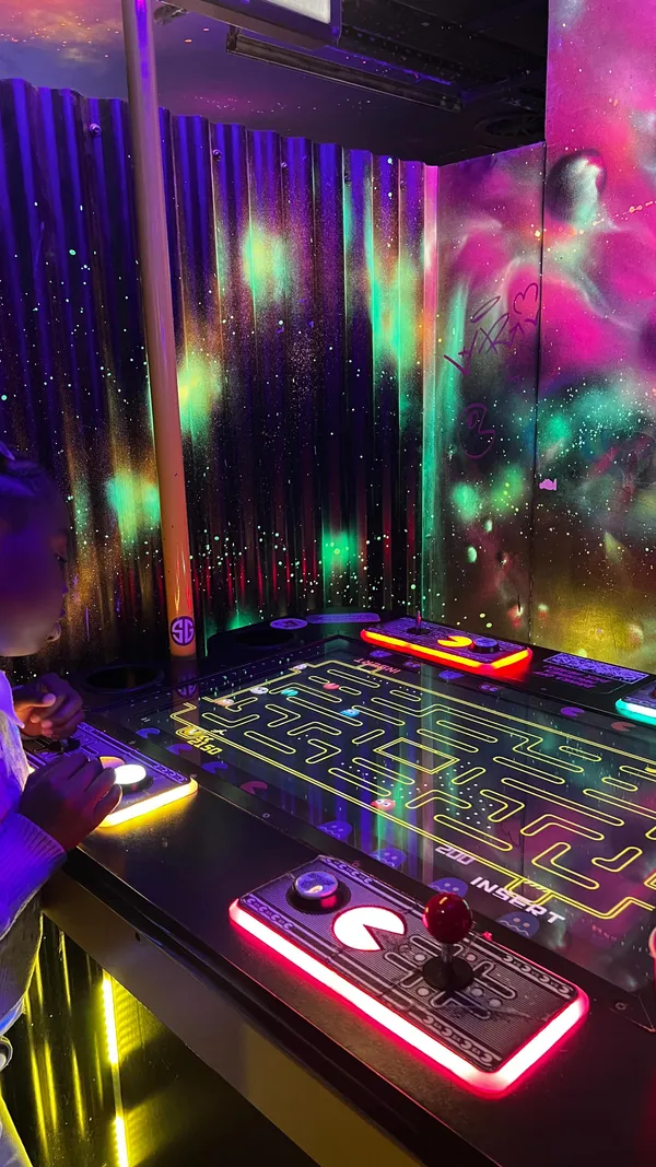 The video is a promotional piece for NQ64, a retro gaming arcade in London, highlighting its family-friendly 'Shorty Sundays'. It follows a person through the neon-lit, graffiti-art-covered venue, showcasing a wide variety of games being played, including dancing machines, Daytona USA, Marvel vs. Street Fighter, Guitar Hero, X-Men pinball, and a tabletop Pac-Man. The video also features a shot of two glowing green soft drinks, emphasizing the kid-friendly options available. The overall focus is on the vibrant, fun, and nostalgic atmosphere of the arcade.