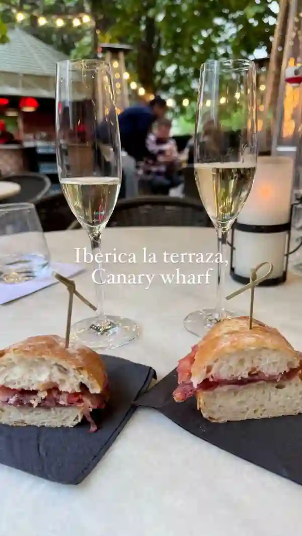 The video showcases Iberica La Terraza, a Spanish restaurant in Canary Wharf, London. It highlights the restaurant's summer terrace with its charming setup, featuring various seating areas, string lights, and heating elements. The food shown includes a selection of cheeses, Spanish ham, and small sandwiches. The drinks shown include champagne. The presentation of the food is simple but appealing. The main focus of the video is to showcase the ambiance and food of the summer terrace. No dietary information is explicitly provided. 